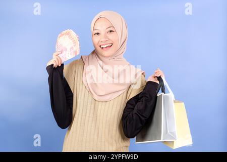 Eine asiatische junge Frau, die Hijab und Pullover trägt, hält Geld Rupiah Cash und Einkaufstaschen, isoliert auf blauem Hintergrund Stockfoto