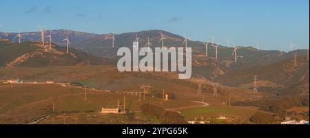 Die sanften grünen Hügel in der Nähe von Tarifa Spanien bieten zahlreiche Windturbinen vor einem Hintergrund von Bergen und einem klaren blauen Himmel mit Stromleitungen und Bauwerken Stockfoto