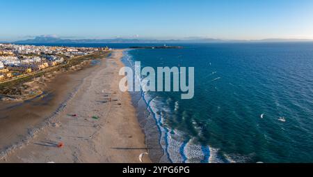 An der Küste von Tarifa gibt es einen Sandstrand, ein pulsierendes blaues Meer und farbenfrohe Kitesurfer. Ein Leuchtturm steht in der Ferne unter klarem Himmel. Stockfoto