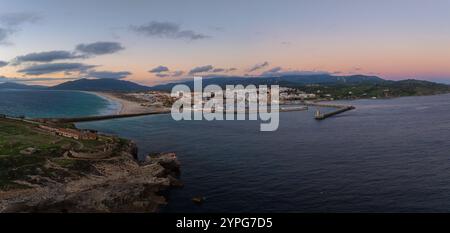 Tarifa, Spanien, ist von oben mit seinen weißen Gebäuden, dem Sandstrand und dem Hafen zu sehen. Der Himmel zeigt Rosa- und Orangetöne, die auf Sonnenuntergang hinweisen. Stockfoto