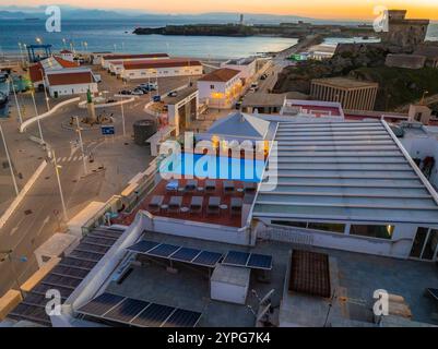 Tarifa, Spanien, ist von oben bei Sonnenuntergang zu sehen, mit einem Dachpool, historischer Struktur, Küste und entferntem Leuchtturm unter einem warmen Licht. Stockfoto