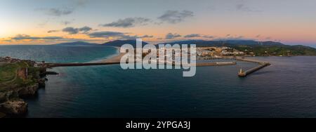 Tarifa, Spanien, ist von oben gesehen, mit einem Sandstrand, Hafen mit Wellenbrecher, weißen Gebäuden und sanften Hügeln unter einem orange-rosa Himmel. Stockfoto