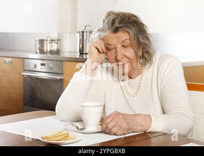 Eine alte Frau sitzt am Küchentisch mit einer Tasse Tee und Keksen Stockfoto