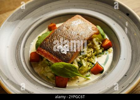 Ein perfekt gegrilltes Lachsfilet, serviert mit geröstetem Gemüse und einer cremigen Sauce auf einem dekorativen Teller. Das Gericht ist mit Spinatblättern garniert, Stockfoto