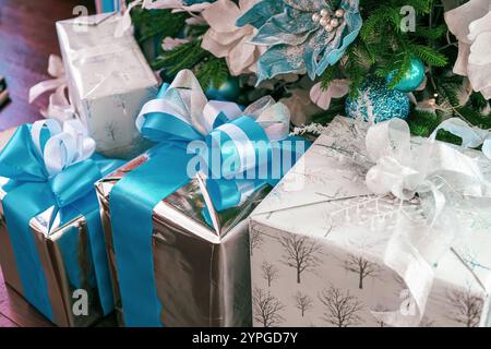 Glänzende Geschenke in Silber und weiß mit blauen Bändern sind unter einem üppigen Weihnachtsbaum mit Ornamenten und Blüten angeordnet. Das f Stockfoto