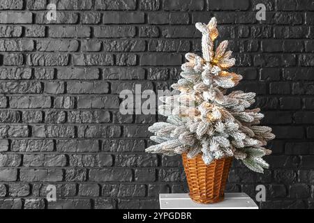 Weihnachtsbaum in einem gewebten Korb, beleuchtet von warmen Lichtern mit Frost, schneebedeckten Zweigen mit Lichtern auf dunklem Backsteinmauerhintergrund. Hintergrundbild, Stockfoto