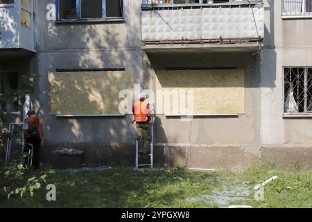 Ein Wohnhaus mit den Folgen eines Raketenangriffs und provisorischen Fensterreparaturen. Ein Arbeiter deckt kaputte Fenster mit Sperrholz ab. Stockfoto