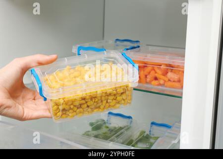 Frau, die Plastikbehälter mit gefrorenem Mais aus dem Kühlschrank nimmt, Nahaufnahme Stockfoto