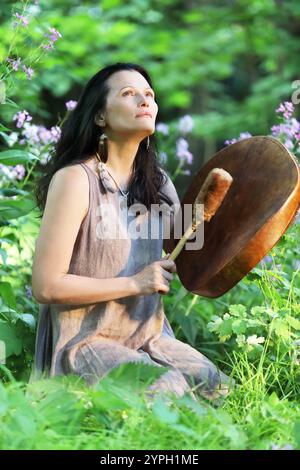 Schamanische Frau spielt auf schamanischer Rahmentrommel bei Sonnenaufgang im Wald auf Hintergrund mit Blättern und Blumen Stockfoto