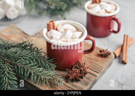 Zwei rote Emailtassen mit heißer Schokolade, Marshmallows und Zimtstangen auf einem Holzschneidebrett mit Tannenzweigen auf dem Hintergrund Stockfoto