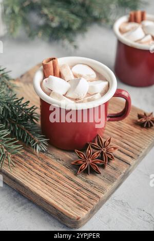 Zwei rote Emailtassen mit heißer Schokolade, Marshmallows und Zimtstangen auf einem Holzschneidebrett mit Tannenzweigen auf dem Hintergrund Stockfoto