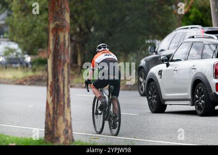Rückansicht eines Mannes in schwarz, orange und weiß, während er mit seinem Fahrrad auf einer zweispurigen Straße neben Autos fährt Stockfoto