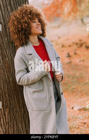 Eine junge Frau mit lockigen Haaren lehnt sich an einen Baum, trägt einen warmen Pullover, lächelt fröhlich im goldenen Herbstlaub und umhergefallenen Blättern Stockfoto