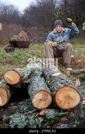 Ein älterer Mann sitzt zwischen Baumstämmen, die er geschnitten hat, während er Mistel über Kopf hält, während er Kuss-Konzept allein einsames weihnachten bläst Stockfoto