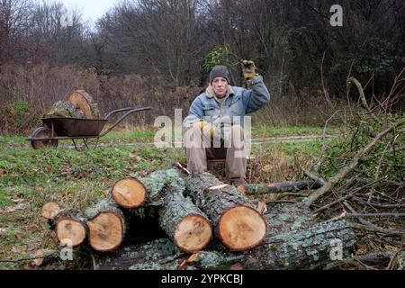 Ein älterer Mann sitzt zwischen Baumstämmen, die er geschnitten hat, während er Mistel über Kopf hält, während er Kuss-Konzept allein einsames weihnachten bläst Stockfoto