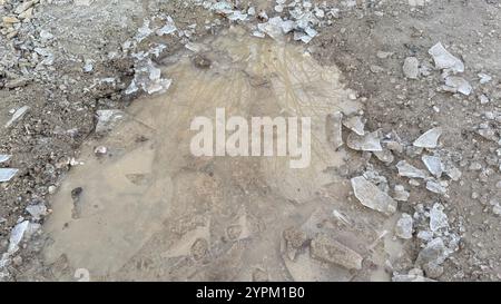 Eine schlammige Pfütze mit gebrochenen Eissplittern, die auf einem unbefestigten Boden verstreut sind. Die Pfütze reflektiert das unscharfe Bild von Ästen. Der Boden ist kompo Stockfoto