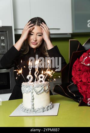 Eine Frau bläst Kerzen auf einem Kuchen mit der Nummer 28. Sie trägt ein schwarzes Kleid und hat ihre Hände auf dem Kopf. Der Kuchen wird auf einen gelegt Stockfoto