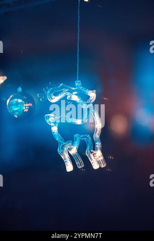 Zauberhafte Weihnachtsszene mit Blauhirsch, Glocke und baumförmigen Ornamenten, die vor Funkeln leuchten. Die bunten Lichter, die sich im Fenster spiegeln Stockfoto