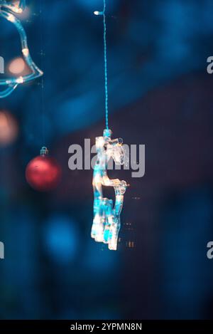 Zauberhafte Weihnachtsszene mit Blauhirsch, Glocke und baumförmigen Ornamenten, die vor Funkeln leuchten. Die bunten Lichter, die sich im Fenster spiegeln Stockfoto