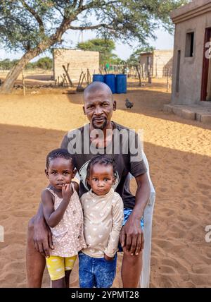 afrikanische Dorffamilie, Vater mit zwei Kindern mit Zöpfen, saß auf einem Stuhl im Hof Stockfoto