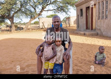 afrikanische Dorffamilie, Porträt glücklicher Vater, der mit zwei Kindern spielt Mädchen mit Zöpfen, sitzt auf einem Stuhl im Garten Junge im Hintergrund im Sand Stockfoto