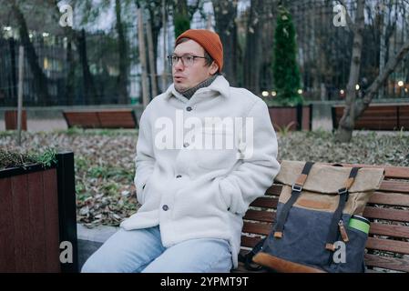 Junger Mann mit Brille und Winterkleidung, der an einem kalten Herbsttag in einem Park auf einer Bank sitzt, seine Hände in den Taschen hält und nachdenklich wegblickt, mit einem Rucksack neben ihm Stockfoto