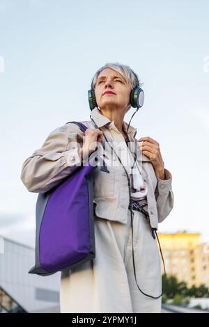 Durchdachte Reife Frau mit grauem Haar steht draußen in der Stadt, trägt Kopfhörer und beige Jacke, schaut Distanz und reflektiert das Leben auf Whit Stockfoto