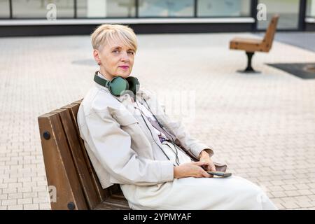 Schöne ältere Frau mit kurzen Haaren, die Kopfhörer trägt und das Smartphone benutzt, während sie draußen in der Stadt auf der Bank sitzt, aktive Lifestyle für ältere Werbespots Stockfoto