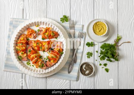 Uunimakkara, gebacken im Ofen Würstchen mit Käse, Tomaten, Koriander, Senf in Auflaufform auf weißem Holztisch mit Gabeln, finnischer Retro-Rezept Stockfoto