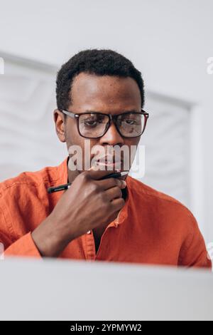 Junger Geschäftsmann mit Brille und orangefarbenem Hemd, einem Stift am Mund und einem Blick auf seinen Laptop, während er von zu Hause aus arbeitet Stockfoto