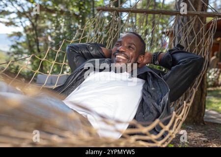 Unbeschwerter afroamerikanischer Mann, der einen Glamping-Kurzurlaub aus der Stadt genießt, während er sich in einer Hängematte entspannt. Stockfoto