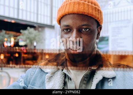 Ein stilvoller junger Mann mit orangefarbener Mütze, Denim-Jacke und Rollkragen posiert selbstbewusst in einer verschwommenen urbanen Umgebung mit warmen Hintergrundlichtern Stockfoto