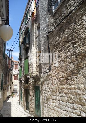 Eine enge Gasse schlängelt sich durch ein historisches Steinviertel mit grünen Fensterläden und Balkonen. Sonnenlicht filtert durch die Gebäude und wirft Schatten Stockfoto