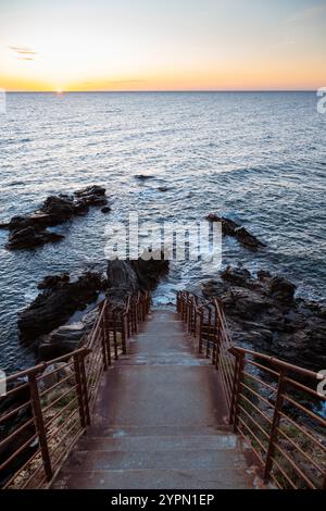 Sonnenaufgang über einer Bucht mit zerklüfteten Felsen, Betontreppe und rostigem Geländer an der Mittelmeerküste in der Nähe von Collioure, Cote Vermeille, Okzitanien Stockfoto