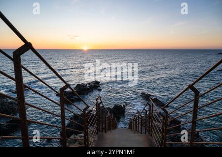 Sonnenaufgang über einer Bucht mit zerklüfteten Felsen, Betontreppe und rostigem Geländer an der Mittelmeerküste in der Nähe von Collioure, Cote Vermeille, Okzitanien Stockfoto