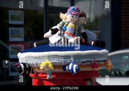 Schrulliger, farbenfroher Strickbezug aus Wolle mit Meeresmotiv und gehäkeltem Schwimmer, Surfer und Fisch schmücken eine traditionelle britische rote Säule. UK. Stockfoto