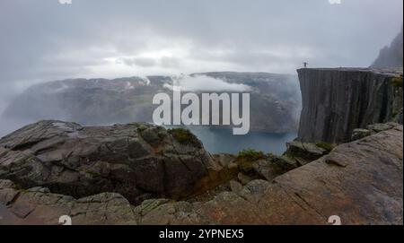 Eine Person steht am Rande einer felsigen Klippe mit Blick auf eine nebelige Fjordlandschaft. Stockfoto