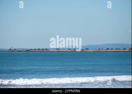 Der lange felsige Pier erstreckt sich unter klarem Himmel in das pulsierende blaue Meer, an dessen Basis Wellen brechen. Stockfoto