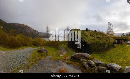 Eine rustikale Hütte mit einem grasbewachsenen Dach befindet sich in einer malerischen Landschaft in der Nähe eines Sees, umgeben von Hügeln und Bäumen. Der Himmel ist bedeckt und schafft eine Ruhe Stockfoto