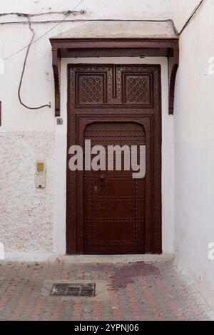 Handgefertigte traditionelle marokkanische Tür mit komplexen geometrischen Mustern und Schnitzereien in einer weißen Gasse. Stockfoto