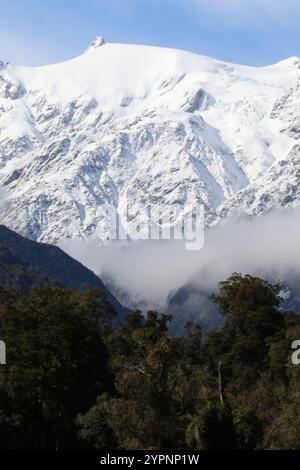 Majestätische schneebedeckte Gipfel erheben sich über üppige Wälder, mit nebeligen Wolken, die durch die Bergtäler treiben. Stockfoto