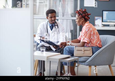 Porträt des afroamerikanischen Arztes in voller Länge, der Laborkittel trägt und ein Klemmbrett hält, während er die Behandlung mit der jungen Frau während der Beratung in der medizinischen Klinik bespricht Stockfoto