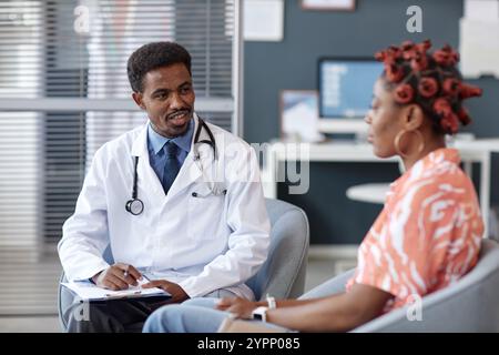 Porträt eines lächelnden afroamerikanischen Arztes, der Labormantel und Stethoskop trägt und mit einer jungen Frau während der Sprechstunde in der Klinik spricht Stockfoto