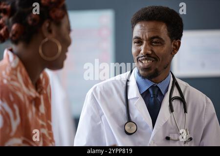 Nahaufnahme Porträt eines freundlichen afroamerikanischen Arztes, der Laborkittel trägt und lächelt, während er der jungen afroamerikanischen Frau während der Konsultation in der Klinik aufmerksam zuhört, Kopierraum Stockfoto
