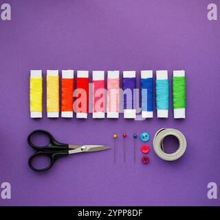 Zusammensetzung von Nähzubehör auf violettem Hintergrund. Bunte Nähgarne, Schere, Nadeln, Knöpfe und Nähzentimeter. Stockfoto