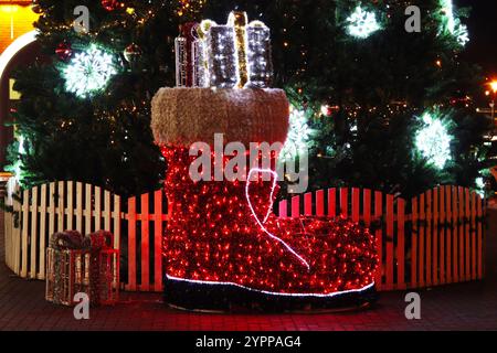 Großer dekorativer Weihnachtsmann-Stiefel mit Girlanden auf dem Hintergrund eines Weihnachtsbaumes, Neujahrsbaum mit leuchtenden Dekorationen. Stockfoto