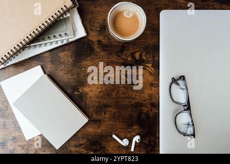 Minimaler Arbeitsplatz mit Kaffeetasse, Spiralnotizbüchern, leerem Papier, Brillen, schnurlosen Ohrstöpseln, und Holzschreibtisch - moderne, stilvolle Einrichtung. Draufsicht. Stockfoto