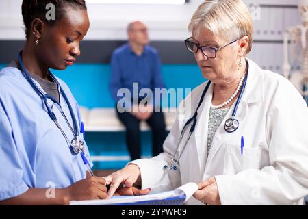 Nahaufnahme eines älteren Arztes, der mit afroamerikanischer Krankenschwester über die Patientenversorgung diskutiert. Frau in blauen Schutteln hält die Zwischenablage und prüft medizinische Daten mit einem leitenden Arzt, während der alte Patient hinter ihnen wartet. Stockfoto