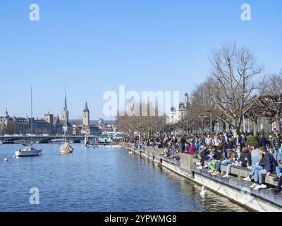 Menschen genießen das erste Frühlingssonnenlicht an der beliebten Wanderpromenade am See in Zürich, Schweiz. Blick auf das Stadtzentrum Stockfoto