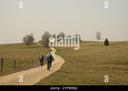 An einem schönen Herbsttag fahren zwei Mountainbiker auf einer Schotterstraße durch eine Wiese zu einem Bauernhof im Schweizer Juragebirge Stockfoto